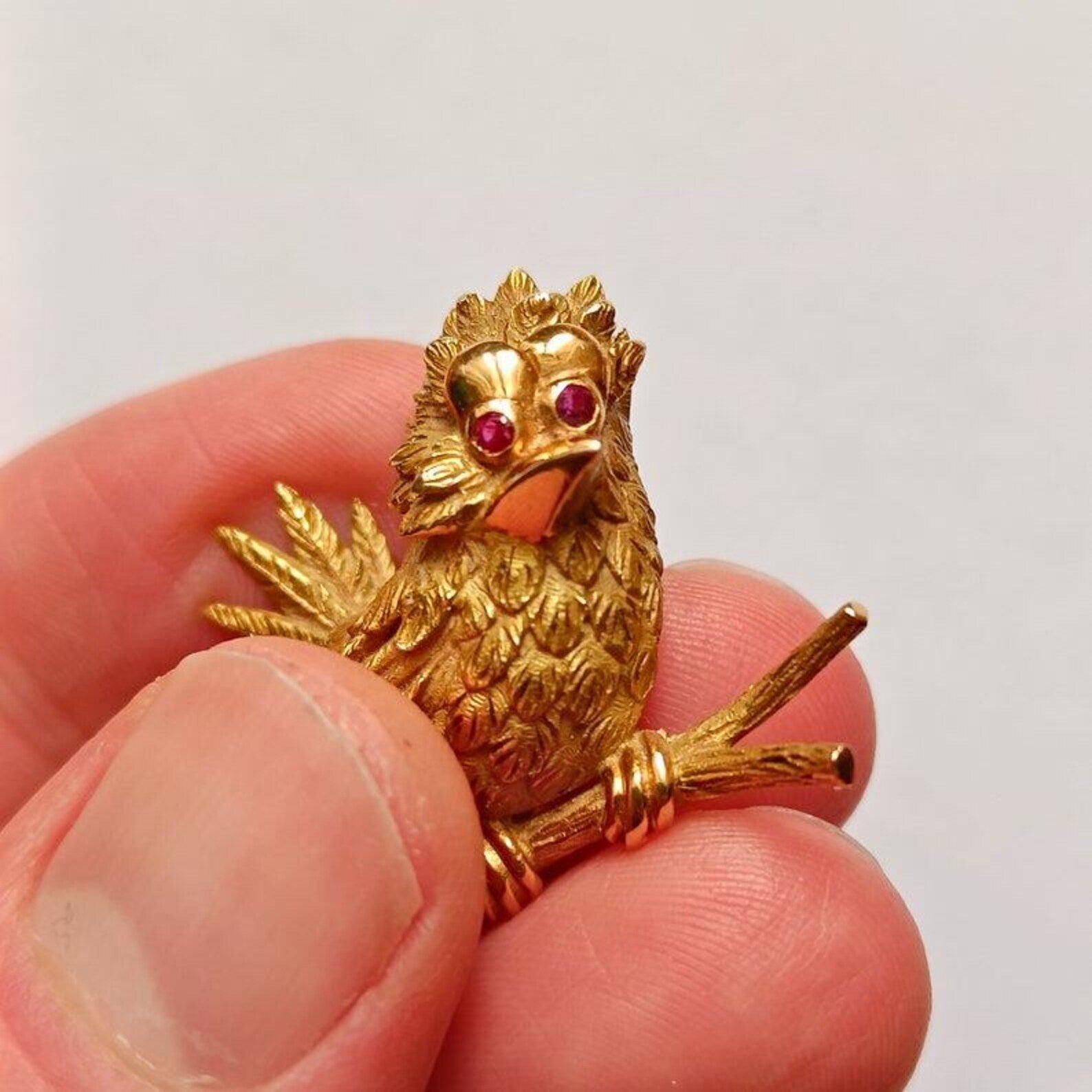 Gold bird-shaped brooch with red eyes held between fingers against a white background